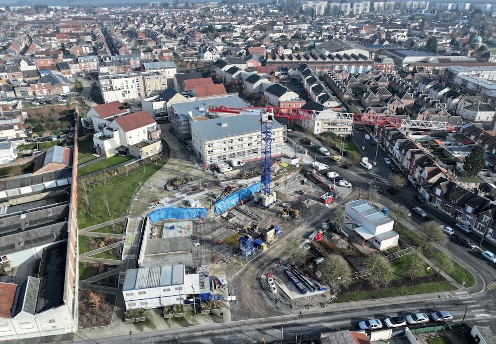 Une vue aérienne du quartier en travaux. Une grande grue est entourée de matériel de construction. Des premiers bâtiments et espaces verts sont visibles, entourés par le reste des quartiers déjà existants. 